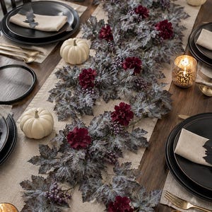 Peut inclure: Une table dressée avec des assiettes noires, des couverts dorés et des serviettes beiges. Une guirlande de feuilles grises, de fleurs bordeaux et de baies violettes orne le centre. Des citrouilles blanches et des bougies allumées complètent le thème automnal.