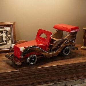 May include: A decorative wooden toy car featuring a red hood, red roof, and dark brown stained wood body with four black wheels. The car is positioned on a wooden mantelpiece alongside a framed photograph, a gold-toned clock, and two books.