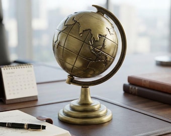 May include: A metallic bronze-toned globe featuring a raised relief map of the continents and a grid of latitude and longitude lines. The globe is mounted on a circular pedestal base with a curved arm that supports the sphere at the poles.