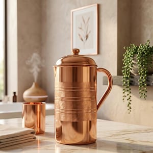 May include: A polished copper water pitcher with a matching cylindrical cup. The pitcher features a domed lid, a curved handle, and horizontal bands of textured, brushed metal finish contrasting with the smooth, reflective copper surface.