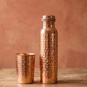 Puede incluir: Una botella de agua de cobre pulido con textura martillada y un vaso de cobre a juego. La botella presenta un cuerpo cilíndrico con un cuello estrecho y una tapa circular plana. Ambos artículos muestran un acabado metálico reflectante.