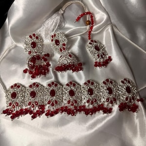May include: A jewelry set consisting of a silver-toned choker necklace, two bell-shaped jhumka earrings, and a maang tikka headpiece. Each piece features intricate silver metalwork, central red oval stones, and dangling red crystal beads.