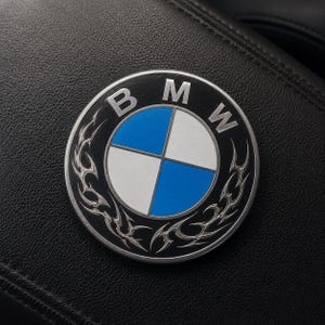 Puede incluir: Un emblema circular de BMW con el icónico diseño de cuadrantes azul y blanco. El anillo exterior negro muestra las letras BMW en plata y está decorado con intrincados y afilados patrones de llamas tribales plateadas en el borde inferior.