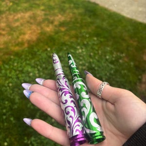Puede incluir: Dos objetos metálicos cilíndricos con forma de casquillos de bala, uno de color morado vibrante y el otro de color verde brillante. Ambos presentan intrincados patrones de volutas plateadas grabados en toda su superficie.