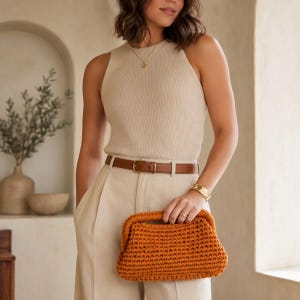 Può includere: Una borsa a mano all'uncinetto color arancio bruciato con una struttura rigida e un manico integrato. La borsa presenta un motivo a maglia denso e uniforme con una silhouette curva, indossata davanti a un top beige smanicato a coste.