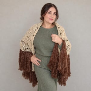 Puede incluir: Un chal de ganchillo beige con un patrón calado en la parte superior y largos flecos marrones densos en el borde inferior. El chal está drapeado sobre los hombros, mostrando el contraste entre las texturas claras y oscuras.