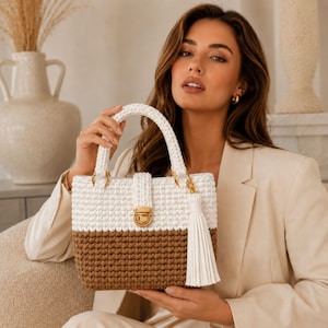 Può includere: Una borsa all'uncinetto bicolore con una sezione superiore bianca e una sezione inferiore marrone. La borsa include un manico superiore, una chiusura a pressione in metallo dorato e una lunga nappa bianca attaccata a un anello dorato sul lato.