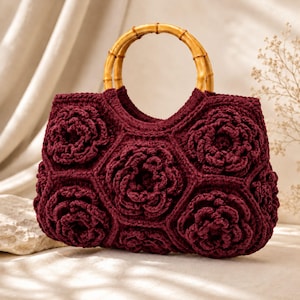 May include: A deep burgundy crochet handbag featuring a series of interconnected hexagonal panels, each adorned with a prominent three-dimensional floral motif. The bag is equipped with two rounded, natural-colored bamboo handles.