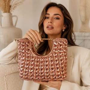 May include: A metallic rose gold handbag with a textured, crocheted surface and a rigid, U-shaped top opening. The bag features a shiny, reflective finish and a structured, rectangular silhouette with a gold-toned metal handle integrated into the top.