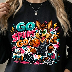 May include: A black long-sleeve sweatshirt featuring a colorful graphic of a cartoon coyote playing basketball. The design includes the text "GO SPURS GO" in teal, pink, and orange, with a city skyline, stars, and lightning bolts in the background.
