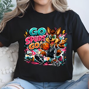 Puede incluir: Una camiseta negra con un gr&aacute;fico colorido de un coyote de dibujos animados jugando al baloncesto. El dise&ntilde;o incluye el texto "GO SPURS GO" en letras llamativas y vibrantes, junto con una canasta de baloncesto, un horizonte urbano y salpicaduras brillantes de rosa, azul y amarillo.