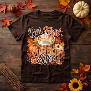 May include: A dark brown short-sleeve t-shirt featuring a graphic print of a coffee cup with whipped cream, surrounded by pumpkins, cinnamon sticks, and orange slices. The text reads But First Pumpkin Spice in a decorative orange and white font.