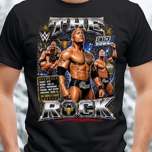 May include: A black t-shirt featuring a graphic of The Rock with the text THE ROCK, WWE, and SMACKDOWN. The design includes a stats box listing height 6'5", weight 260 lb, style powerhouse, finisher rock bottom, hometown Miami, FL, and The People's Champ.