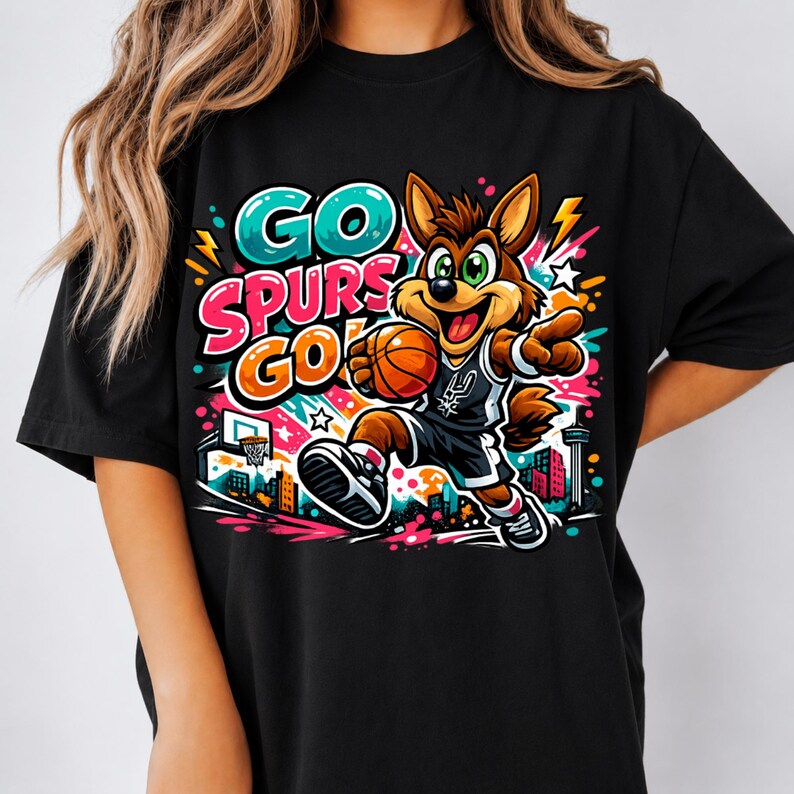 Puede incluir: Una camiseta negra con un gr&aacute;fico colorido de un coyote de dibujos animados jugando al baloncesto. El dise&ntilde;o incluye el texto GO SPURS GO en letras llamativas y vibrantes, junto con estrellas, rayos y un horizonte urbano de fondo.