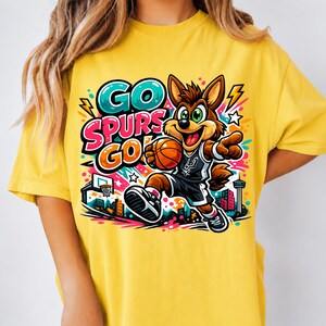 Puede incluir: Una camiseta amarilla con un coyote de dibujos animados que lleva una camiseta de baloncesto y sostiene un bal&oacute;n. El gr&aacute;fico incluye el texto "GO SPURS GO" en letras llamativas y coloridas, junto a estrellas, rayos y una ciudad.