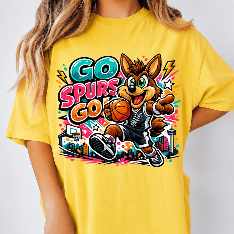 Puede incluir: Una camiseta amarilla con un coyote de dibujos animados que lleva una camiseta de baloncesto y sostiene un bal&oacute;n. El gr&aacute;fico incluye el texto "GO SPURS GO" en letras llamativas y coloridas, junto a estrellas, rayos y una ciudad.