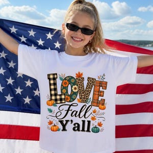 May include: A white short-sleeved t-shirt featuring a graphic print with the text LOVE Fall Y'ALL. The letters are decorated with black and white buffalo plaid, floral, leopard print, and orange plaid patterns, accented by pumpkins and leaves.