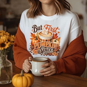 May include: A white crewneck sweatshirt featuring a graphic print of a coffee cup with whipped cream, pumpkins, orange slices, and cinnamon sticks. The text reads But First PUMPKIN Spice in brown and orange script and block lettering.