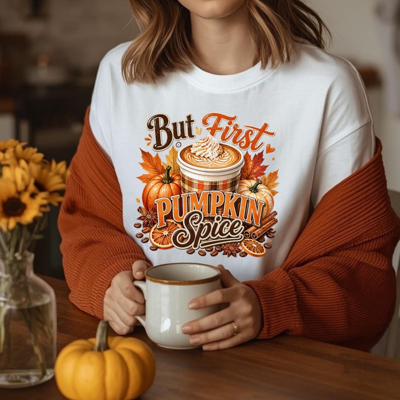 May include: A white crewneck sweatshirt featuring a graphic print of a coffee cup with whipped cream, pumpkins, orange slices, and cinnamon sticks. The text reads But First PUMPKIN Spice in brown and orange script and block lettering.