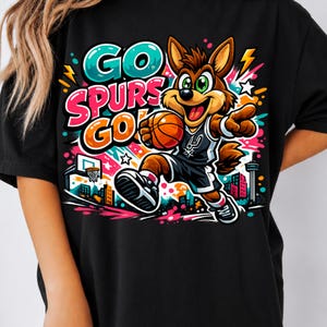Puede incluir: Una camiseta negra con un gr&aacute;fico vibrante de un coyote de dibujos animados jugando al baloncesto. El dise&ntilde;o incluye el texto "GO SPURS GO!" en letras llamativas y coloridas, rodeado de estrellas, rayos y un horizonte urbano estilizado.