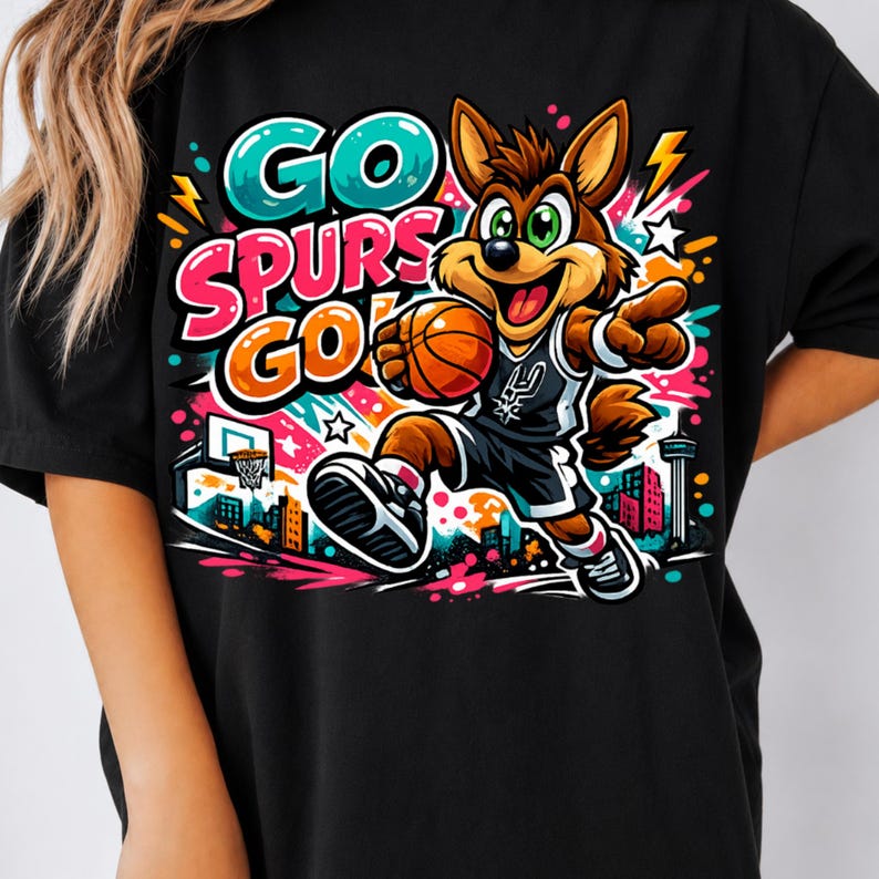 Puede incluir: Una camiseta negra con un gr&aacute;fico vibrante de un coyote de dibujos animados jugando al baloncesto. El dise&ntilde;o incluye el texto "GO SPURS GO!" en letras llamativas y coloridas, rodeado de estrellas, rayos y un horizonte urbano estilizado.