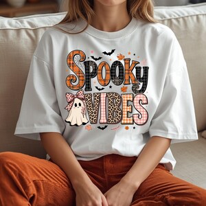 May include: A white short-sleeved t-shirt featuring the text "SPOOKY VIBES" in a mix of orange plaid, black plaid, and leopard print patterns. A small ghost wearing a pink polka-dot bow is positioned below the text, surrounded by bats and leaves.