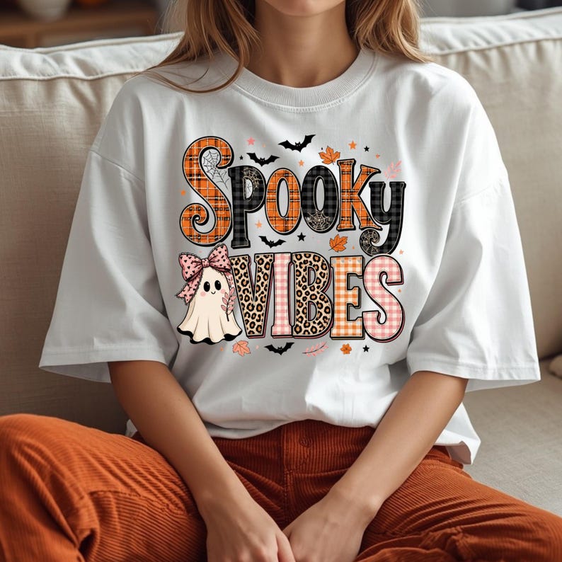 May include: A white short-sleeved t-shirt featuring the text "SPOOKY VIBES" in a mix of orange plaid, black plaid, and leopard print patterns. A small ghost wearing a pink polka-dot bow is positioned below the text, surrounded by bats and leaves.