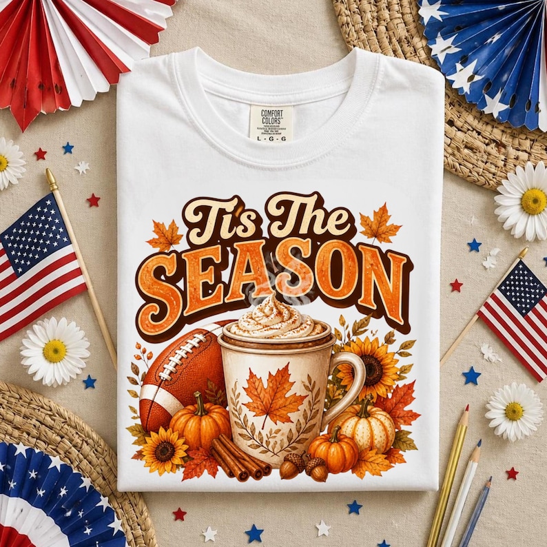 May include: A white t-shirt featuring a graphic with the text 'Tis The Season' in brown and orange. The design includes a football, a mug of coffee with whipped cream, pumpkins, sunflowers, and autumn leaves in shades of orange and brown.