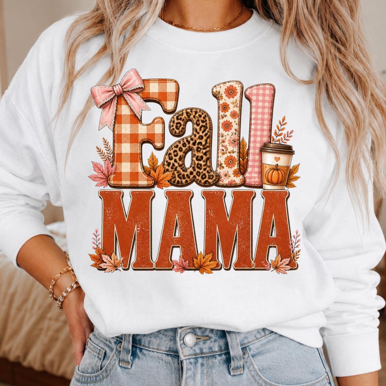 May include: A white long-sleeve sweatshirt featuring a graphic print with the text Fall MAMA. The word Fall is styled with orange plaid, leopard print, floral patterns, and a pink bow. A coffee cup with a pumpkin icon sits next to the text.