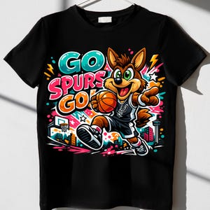 Puede incluir: Una camiseta negra de manga corta con un gr&aacute;fico colorido de un coyote de dibujos animados jugando al baloncesto. El dise&ntilde;o incluye el texto GO SPURS GO en letras llamativas y vibrantes, junto a una canasta, una ciudad y manchas de color.