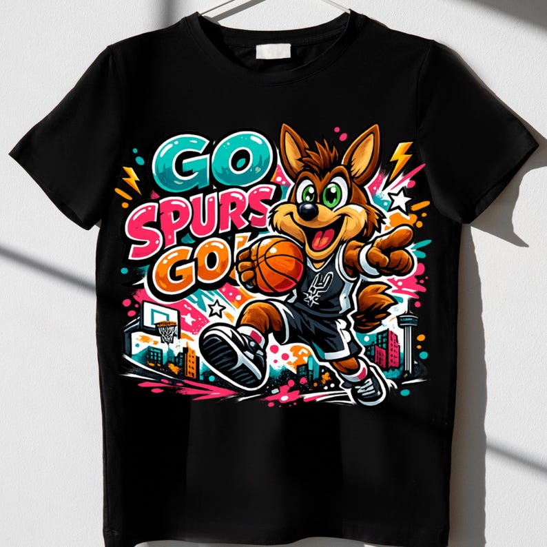Puede incluir: Una camiseta negra de manga corta con un gr&aacute;fico colorido de un coyote de dibujos animados jugando al baloncesto. El dise&ntilde;o incluye el texto GO SPURS GO en letras llamativas y vibrantes, junto a una canasta, una ciudad y manchas de color.