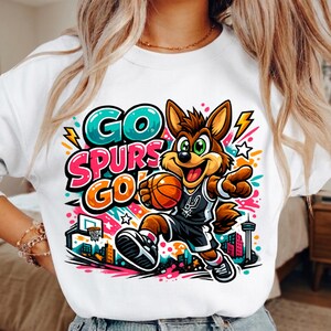 Puede incluir: Una sudadera blanca con un gr&aacute;fico colorido de un coyote de dibujos animados jugando al baloncesto. El texto "GO SPURS GO" est&aacute; impreso en letras llamativas y vibrantes. El dise&ntilde;o incluye una canasta, una ciudad y formas abstractas.