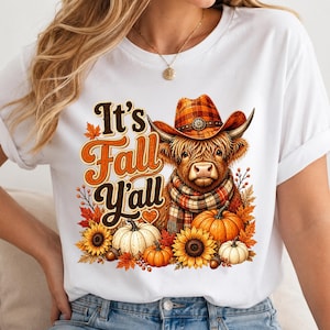 May include: A white t-shirt featuring a graphic of a Highland cow wearing a plaid cowboy hat and scarf. The text reads It's Fall Y'all. The design includes orange and white pumpkins, sunflowers, and autumn leaves in shades of orange and brown.