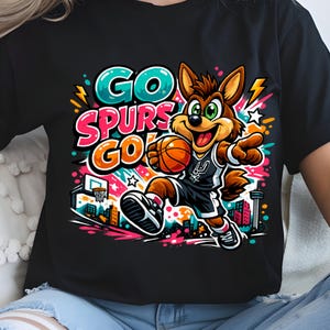 Puede incluir: Una camiseta negra con un gr&aacute;fico colorido de un coyote de dibujos animados jugando al baloncesto. El texto GO SPURS GO est&aacute; impreso en letras grandes y negritas. El dise&ntilde;o incluye un horizonte urbano, una canasta y salpicaduras de color.