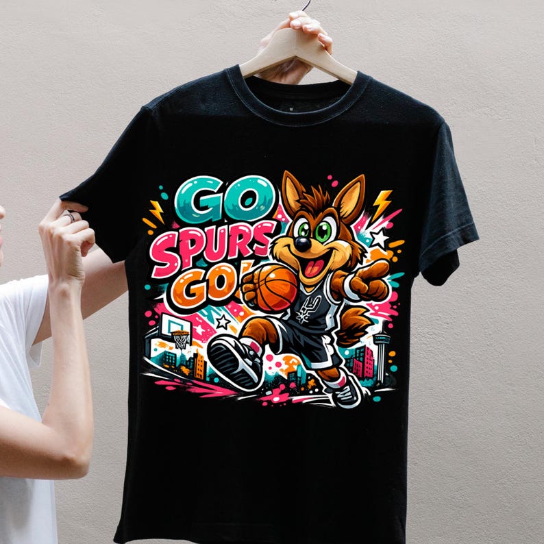 Puede incluir: Una camiseta negra con un gr&aacute;fico colorido de un coyote de dibujos animados jugando al baloncesto. El dise&ntilde;o incluye el texto GO SPURS GO en letras llamativas y vibrantes, junto con estrellas, rayos y una canasta de baloncesto.