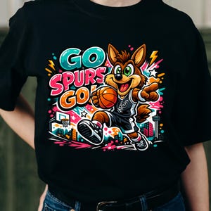 Puede incluir: Una camiseta negra con un gr&aacute;fico colorido de un coyote de dibujos animados con una camiseta de baloncesto, sosteniendo un bal&oacute;n y se&ntilde;alando. El texto GO SPURS GO aparece en letras vibrantes sobre un fondo de formas abstractas y un horizonte urbano.