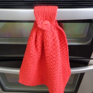 May include: Red knitted oven mitt with a red button. The mitt is hanging on a silver oven door.
