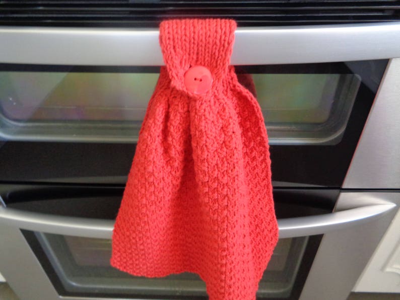 May include: Red knitted oven mitt with a red button. The mitt is hanging on a silver oven door.