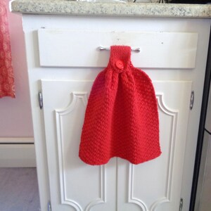 May include: A red knitted dish towel with a red button hanging on a white cabinet door.