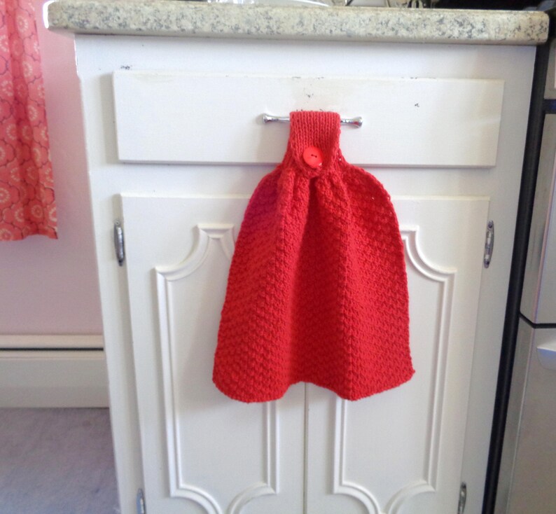 May include: A red knitted dish towel with a red button hanging on a white cabinet door.