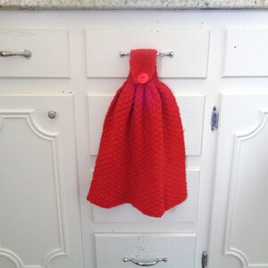May include: A red knitted dish towel with a button hanging on a white kitchen cabinet door.
