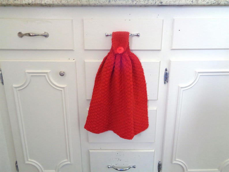 May include: A red knitted dish towel with a button hanging on a white kitchen cabinet door.