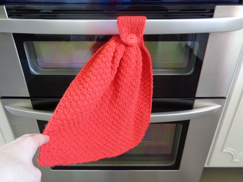 May include: A red knitted oven mitt with a button on the top, hanging on a silver oven door.