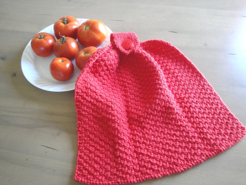 May include: Red knitted dish towel with a button and a textured pattern. The towel is hanging on a white plate with red tomatoes.