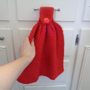 May include: A red knitted dish towel with a button and a metal hook. The towel is hanging on a white cabinet door.