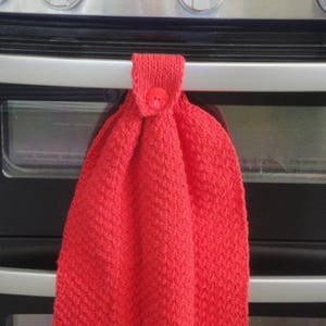 May include: Red knitted oven mitt with a red button hanging on a silver oven door.
