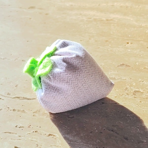 May include: A small, lavender-colored sachet with a woven pattern and a bright green, ruffled closure. The sachet is filled and tied, casting a shadow on a light-colored surface.