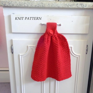 May include: Red knitted dish towel with a button hanging on a hook. The towel is hanging on a white cabinet door with the words "KNIT PATTERN" written on the cabinet door.