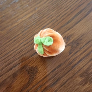 May include: A small, orange velvet pouch with a green bow on top. The pouch is round and has a soft texture. The bow is a lighter shade of green. The pouch is sitting on a wooden surface.