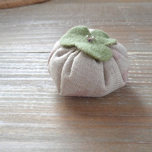 Puede incluir: Un pequeño cojín de alfileres redondo y beige de tela con una hoja verde de fieltro en la parte superior. El cojín está relleno de relleno y tiene una superficie ligeramente texturizada.